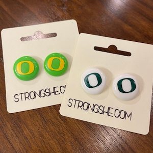 May include: Two pairs of earrings featuring the letter "O" in green and yellow on a white background. The earrings are packaged on white cards with the text "Strongshe.com".