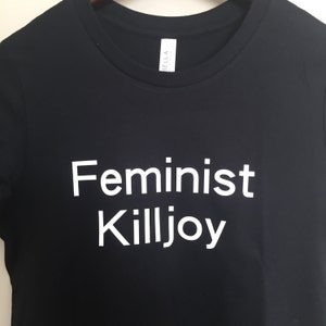 May include: Black t-shirt with the words "Feminist Killjoy" printed in white. The shirt has a crew neck and short sleeves. The Bella brand tag is visible on the upper right side.