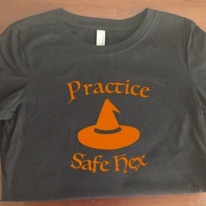 Puede incluir: Camiseta negra con texto naranja que dice "Practice Safe Hex" y un gráfico de sombrero de bruja naranja.