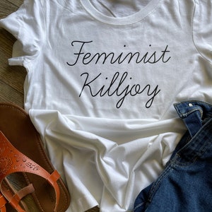 May include: White t-shirt with the text "Feminist Killjoy" printed in black cursive font.