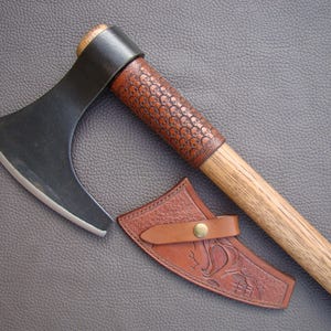 May include: A handcrafted axe with a dark blade and a wooden handle. The handle is wrapped with brown leather, and a matching leather sheath is included. The axe blade is approximately 6 inches long.
