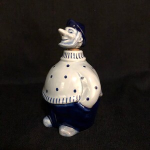 May include: A blue and white ceramic bottle stopper shaped like a man wearing a blue hat and sweater with white polka dots. The man has a large nose and a smiling face.