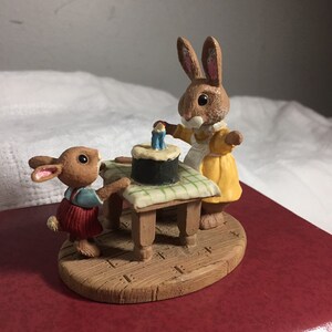 Pode incluir: Dois coelhos marrons, um com um vestido amarelo e o outro com um traje vermelho e azul, est&atilde;o celebrando com um bolo preto e branco com velas azuis em uma mesa de madeira com uma toalha de mesa quadriculada verde e branca.