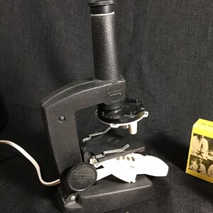 May include: A black microscope with a white stage and a black and white knob. The microscope is plugged in and has a white cord. The microscope is on a black surface. There is a book and a box with pictures of a microscope on the surface.