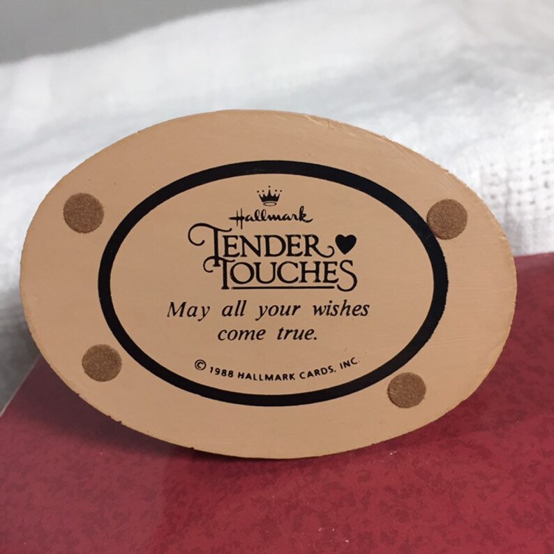 Pode incluir: Uma base de figurinha de cer&acirc;mica ovalada de cor bege com um logotipo preto Hallmark Tender Touches e o texto "May all your wishes come true." &copy; 1988 Hallmark Cards, Inc.