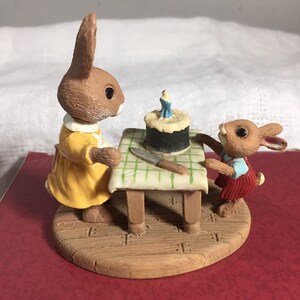 Pode incluir: Duas estatuetas de coelhos marrons, uma com um vestido amarelo e a outra com um vestido vermelho, est&atilde;o em volta de uma mesa pequena com um bolo de anivers&aacute;rio e uma faca. O bolo tem uma &uacute;nica vela acesa.