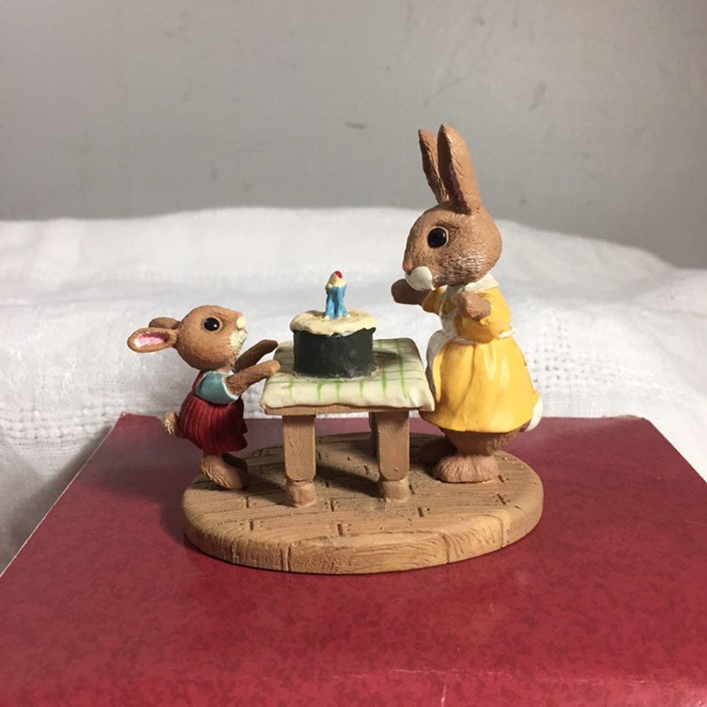 Pode incluir: Uma figura de dois coelhos marrons, um vestindo um vestido amarelo e o outro vestindo um vestido vermelho e azul, em p&eacute; em torno de uma mesa pequena com um bolo de anivers&aacute;rio em cima.