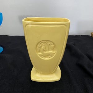 May include: A pale yellow ceramic vase with a rectangular shape and a raised design of two horses in a circle on the front.