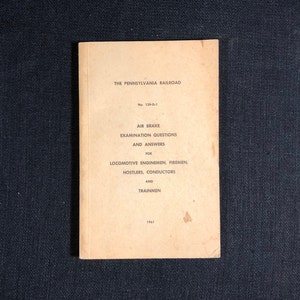 May include: A tan-colored book with the title "The Pennsylvania Railroad" and the text "Air Brake Examination Questions and Answers for Locomotive Enginemen, Firemen, Hostlers, Conductors and Trainmen". The book was published in 1961.