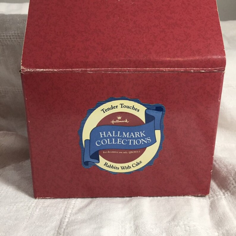 Pode incluir: Caixa de cart&atilde;o vermelha com um adesivo circular branco e azul que diz "Tender Touches Hallmark Collections para uso decorativo apenas QXC973-1 Coelhos com bolo".