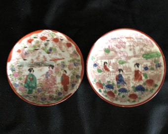 May include: Two small, round, white ceramic plates with a red rim. The plates are decorated with a colorful Japanese-inspired design featuring women in traditional clothing, flowers, and trees.