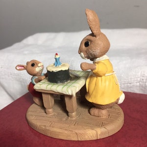 Pode incluir: Uma figurinha de coelho marrom com um vestido amarelo est&aacute; ao lado de uma mesa com um pequeno bolo preto e uma &uacute;nica vela acesa. Uma figurinha de coelho marrom menor com um vestido vermelho olha para o bolo.