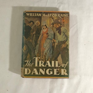 May include: A vintage paperback book cover with a Western theme. The cover features a group of people in a dimly lit room, including a woman in a white dress and a man with a gun. The title of the book is "The Trail of Danger" by William MacLeod Raine.