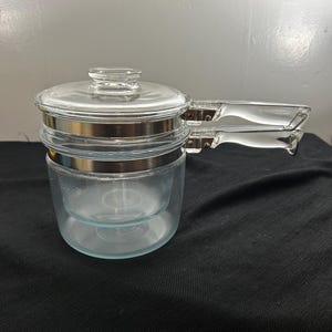 May include: A clear glass double boiler with a glass lid and a clear handle. The double boiler has a silver metal band around the middle and a clear glass handle. The lid has a small glass knob.