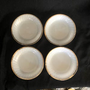 May include: Four white, round saucers with a gold rim and a subtle swirl pattern. The saucers are arranged in a square formation against a dark background, showcasing their elegant design.