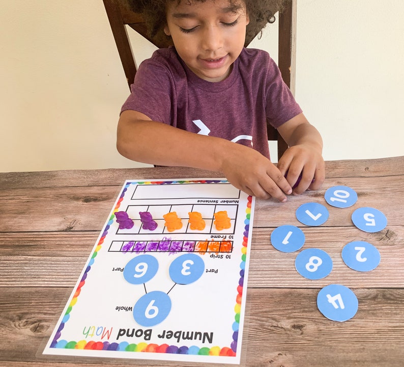 May include: A child is learning about number bonds with a colorful worksheet and blue circles with numbers on them. The worksheet has a number sentence, 10 strip, and 10 frame. The child is using gummy bears to represent the numbers.