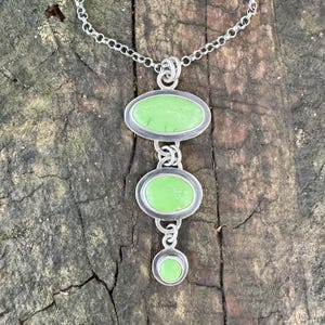 May include: A silver necklace with a pendant featuring three light green oval and round stones. The stones are set in silver bezels and connected by silver links. The necklace is displayed on a wooden surface.