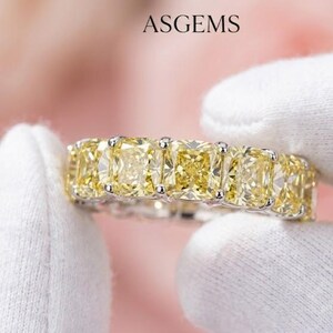 Puede incluir: Una alianza de eternidad plateada con una fila continua de piedras preciosas amarillas de corte radiante engastadas en garras de metal. La palabra ASGEMS está impresa en negro sobre el anillo. El anillo es sostenido por dedos cubiertos con tela blanca.