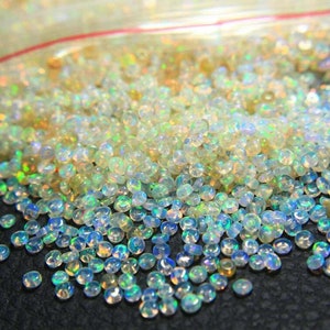 May include: A close-up shot of a pile of small, iridescent opal beads. The beads are mostly clear with flashes of blue, green, and yellow, creating a shimmering effect. They appear to be loose and scattered.
