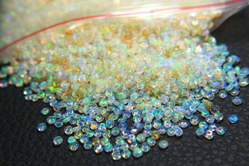 May include: A close-up shot of a pile of small, iridescent opal beads. The beads are mostly clear with flashes of blue, green, and yellow, creating a shimmering effect. They appear to be loose and scattered.