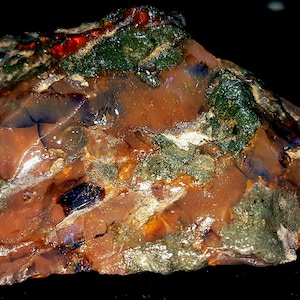 May include: A rough, uncut opal gemstone with a mix of colors. The stone has a reddish-brown base with patches of green and areas that display flashes of red and blue. The surface is textured and uneven, with visible cracks.