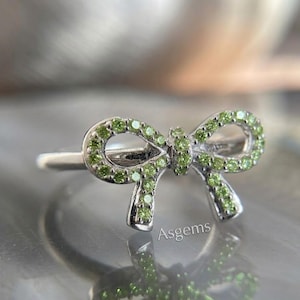 May include: A silver ring in the shape of a bow, encrusted with small, round, light green gemstones. The ring's band is thin and polished, and the bow design is detailed and elegant. The gemstones are evenly spaced, creating a sparkling effect.
