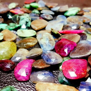 May include: A collection of faceted gemstones in various colors, including red, pink, blue, green, and yellow. The gemstones are scattered across a dark surface, showcasing their different shapes and sizes.