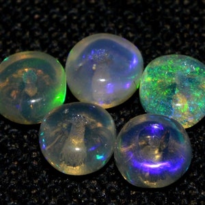 May include: Five iridescent opal gemstones. The opals display a range of colors, including green, blue, and yellow, with a shimmering, translucent quality. The stones are round and polished, reflecting light and creating a play of color.