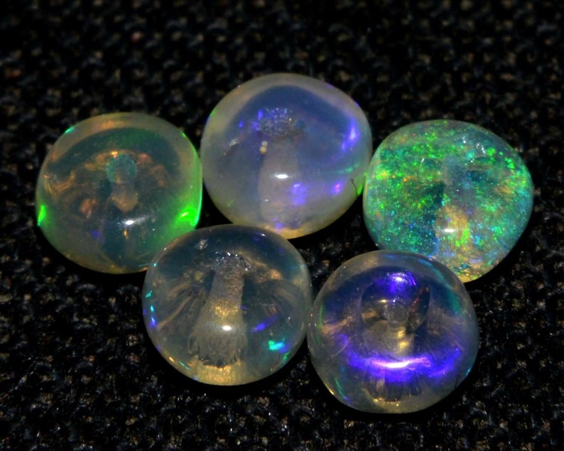 May include: Five iridescent opal gemstones. The opals display a range of colors, including green, blue, and yellow, with a shimmering, translucent quality. The stones are round and polished, reflecting light and creating a play of color.