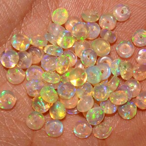 May include: A collection of small, round opal beads with a hole drilled through the center. The beads display a range of colors, including yellow, orange, and green, with iridescent flashes. The beads are scattered across a light-colored surface.