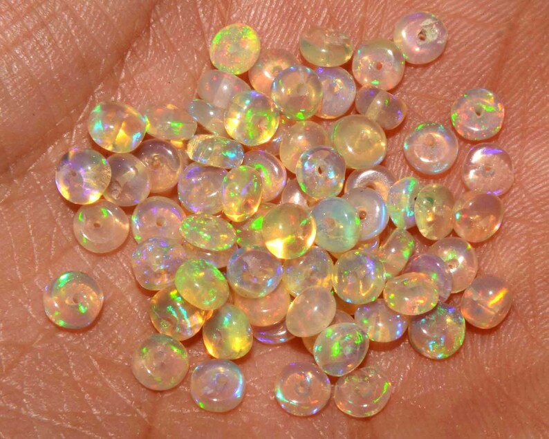 May include: A collection of small, round opal beads with a hole drilled through the center. The beads display a range of colors, including yellow, orange, and green, with iridescent flashes. The beads are scattered across a light-colored surface.