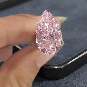 Peut inclure: Une grande pierre précieuse rose en forme de poire montée sur une bague en métal. La pierre présente de nombreuses facettes qui reflètent la lumière, créant un aspect brillant. Le mot asgems est imprimé en bas de l'image.