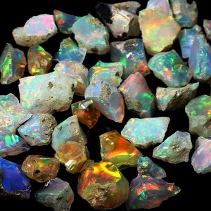 May include: A collection of raw opal gemstones, showcasing a variety of colors including blues, greens, oranges, and yellows. The opals have a rough, natural texture and exhibit iridescent play-of-color. The gemstones are scattered against a dark background.