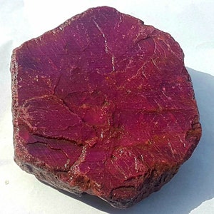 Peut inclure: Gros rubis brut de couleur rouge foncé. La surface de la pierre présente un aspect texturé avec des lignes et des imperfections visibles. Le rubis est de forme irrégulière, mettant en valeur son état naturel et non poli.