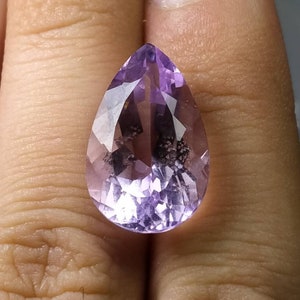 Puede incluir: Una gema facetada en forma de pera, de color p&uacute;rpura claro. La piedra se sostiene en una mano y tiene una superficie pulida con inclusiones internas. El corte de la piedra crea m&uacute;ltiples superficies reflectantes.