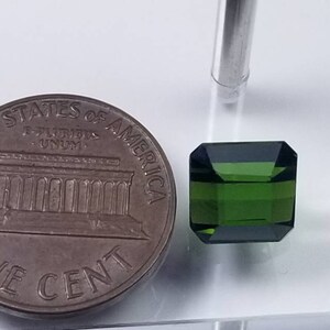 Pode incluir: Uma gema verde esmeralda facetada em forma retangular, ao lado de uma moeda de um centavo. A gema tem uma cor escura e saturada. A moeda &eacute; de cor bronze. A gema mede aproximadamente 0,5 cm.