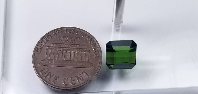 Pode incluir: Uma gema verde esmeralda facetada em forma retangular, ao lado de uma moeda de um centavo. A gema tem uma cor escura e saturada. A moeda &eacute; de cor bronze. A gema mede aproximadamente 0,5 cm.