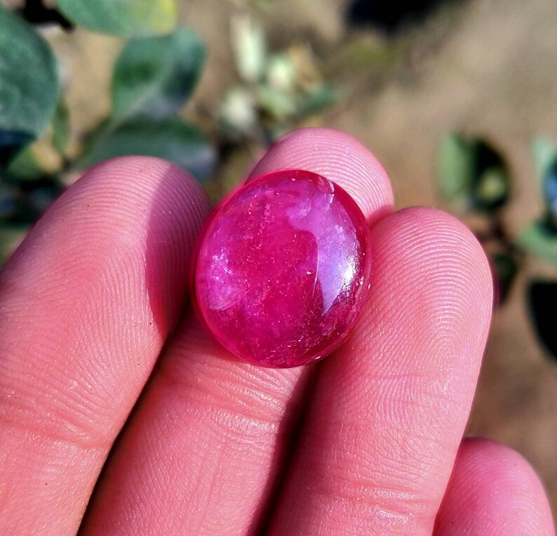 Peut inclure: Une pierre pr&eacute;cieuse de forme ovale, polie, de couleur rouge-rose intense. La surface est lisse et r&eacute;fl&eacute;chissante. La gemme est tenue dans une main, avec un arri&egrave;re-plan flou de feuillage vert.