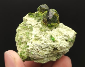 May include: A natural mineral specimen featuring two dark green crystals atop a light green and white rock matrix. The crystals have a faceted appearance, and the matrix has a textured, porous surface. The background is black.