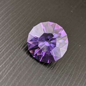 May include: A faceted, round gemstone with a deep purple hue. The gem features a complex, geometric cut that reflects light, creating a sparkling effect. The stone is set against a dark background.