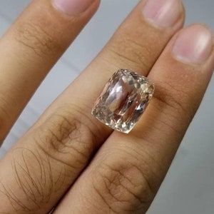 May include: A faceted, rectangular gemstone with a light peach hue. The gem is held between fingers, showcasing its clarity and cut. The stone's facets reflect light, creating a sparkling effect, ideal for jewelry.
