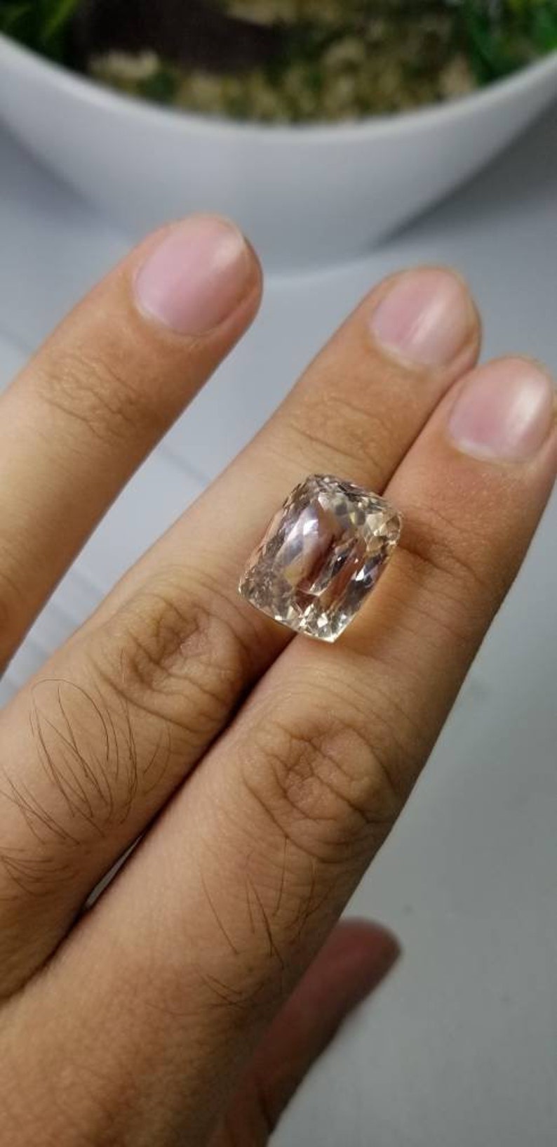 May include: A faceted, rectangular gemstone with a light peach hue. The gem is held between fingers, showcasing its clarity and cut. The stone's facets reflect light, creating a sparkling effect, ideal for jewelry.