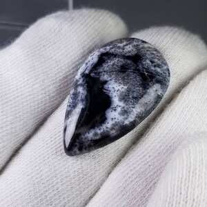 Pode incluir: Uma pedra preciosa em forma de gota, com superf&iacute;cie polida. A pedra apresenta um padr&atilde;o marcante de preto e branco, assemelhando-se a uma paisagem ou design abstrato. A gema &eacute; exibida sobre um fundo de tecido branco.