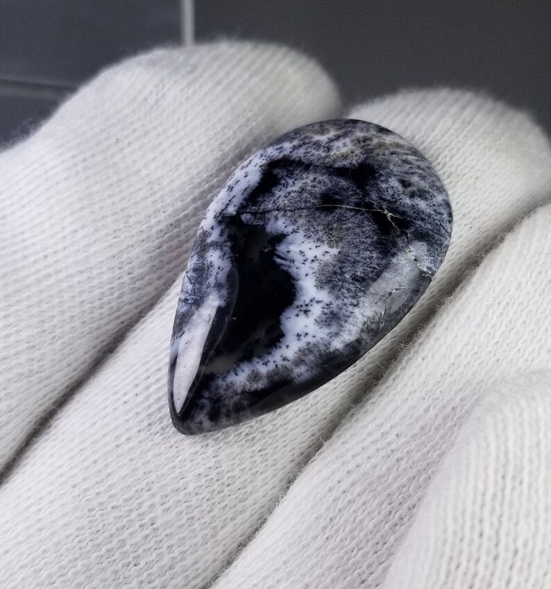 Pode incluir: Uma pedra preciosa em forma de gota, com superf&iacute;cie polida. A pedra apresenta um padr&atilde;o marcante de preto e branco, assemelhando-se a uma paisagem ou design abstrato. A gema &eacute; exibida sobre um fundo de tecido branco.