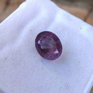 May include: An oval-cut, faceted gemstone with a deep purple hue. The gem is set against a white background, showcasing its clarity and color. The gemstone's facets reflect light, creating a sparkling effect. This gem is suitable for jewelry making.