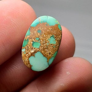 Puede incluir: Una gema ovalada con una base turquesa, que presenta un patr&oacute;n distintivo de matriz marr&oacute;n y dorada. La piedra se sostiene entre los dedos, resaltando su belleza natural y variaciones de color. Ideal para la creaci&oacute;n de joyas.