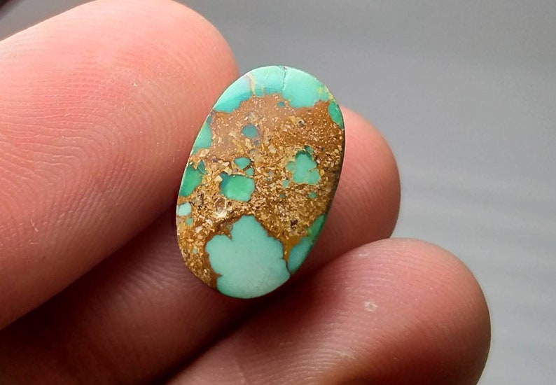 Puede incluir: Una gema ovalada con una base turquesa, que presenta un patr&oacute;n distintivo de matriz marr&oacute;n y dorada. La piedra se sostiene entre los dedos, resaltando su belleza natural y variaciones de color. Ideal para la creaci&oacute;n de joyas.
