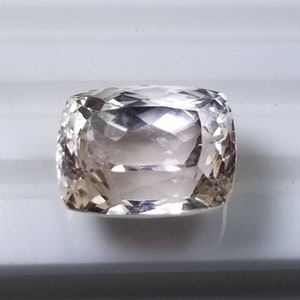 May include: A cushion-cut gemstone with a pale champagne color. The gem has a faceted surface that reflects light, creating a sparkling effect. The stone is set against a white background, highlighting its clarity and brilliance. This gemstone is suitable for jewelry.
