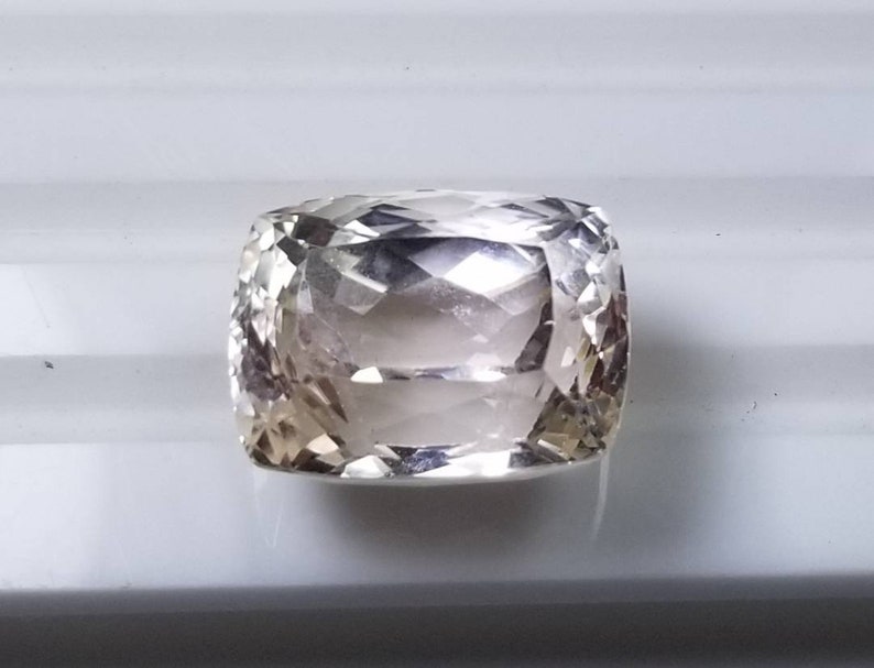 May include: A cushion-cut gemstone with a pale champagne color. The gem has a faceted surface that reflects light, creating a sparkling effect. The stone is set against a white background, highlighting its clarity and brilliance. This gemstone is suitable for jewelry.