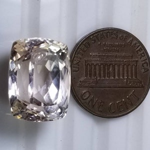 May include: A faceted, light-colored gemstone with a rectangular cushion cut, next to a United States one-cent coin. The gemstone has a translucent appearance and reflects light, showcasing its multiple facets. The coin is bronze-colored and displays the words "United States of America."